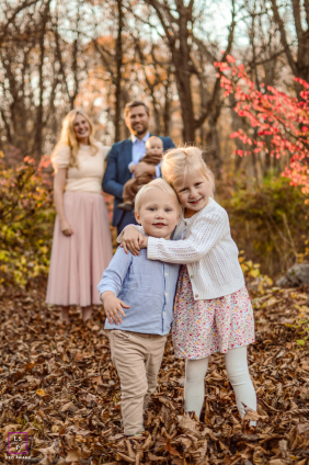 In a customer's backyard, siblings share a warm hug in the foreground, surrounded by colorful fall leaves. The outdoor autumn scene highlights their closeness and the joy of family time together.