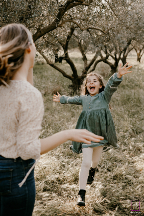 In an olive grove at Campagne, a young girl runs and jumps through the grass, playing joyfully with her mom among the trees in the orchard, energy and laughter filling the scene.