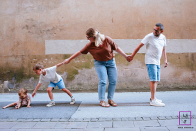 In Bayonne, France, a family walks in single file along the sidewalk: mom, dad, and boy holding hands. The boy reaches back to connect with a seated toddler, emphasizing togetherness and care.