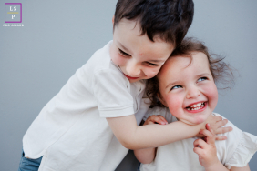 In Paris, toddler brother and sister share a joyful hug, both smiling brightly. The portrait features a clean white background, highlighting their sweet expressions and sibling connection.