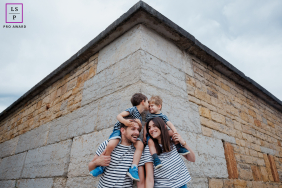In Paris, a family poses for a graphic portrait at the corner of an Old World stone building. Parents carry their kids on their shoulders, captured from a low, wide angle, all smiling brightly.