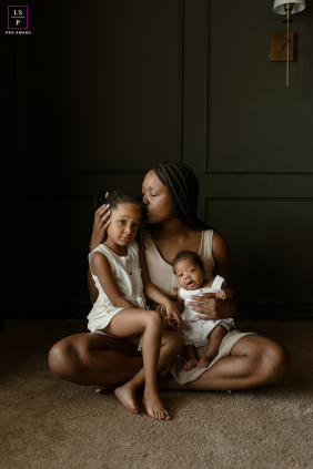 In San Antonio, Texas, a mom sits in her living room with her two daughters on her lap. They are closely nestled together, radiating warmth and affection in their cozy home environment.