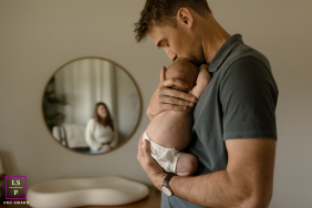 In San Antonio, Texas, a dad gently holds his newborn while the mother's reflection appears in a nearby mirror, capturing both parents' presence and love in a tender family scene.