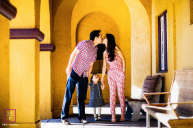 In Burlingame, California, a family portrait shows mom and dad sharing a kiss with their child standing between them, looking up at the kiss, capturing a loving and playful family connection.