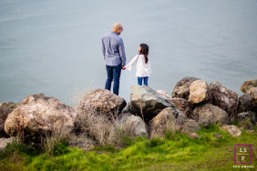 In Emeryville, California, a father and daughter hold hands while standing together on stones by the water. The serene setting highlights their close bond in this heartfelt family portrait.