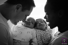In Île-de-France, newborn twins lie cheek to cheek in the center of the frame, with their parents on either side, gently cradling and framing them in a tender black-and-white photograph.