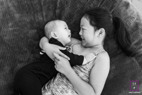 In Santa Barbara, California, siblings lie face to face on a couch, sharing big smiles. This black-and-white, tightly framed photo captures their joyful connection in an intimate, candid scene.