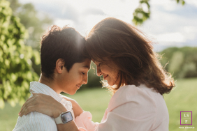In Boston, Massachusetts, a mother and son pose head to head by the trees, warmly lit by the light. Their forms are softly outlined, highlighting their close bond in a peaceful setting.
