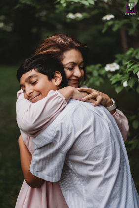 In Boston, Massachusetts, a mom and her son share a warm hug by the trees in a vertical shot. Dappled sunlight filters through the leaves, capturing their close, loving connection.