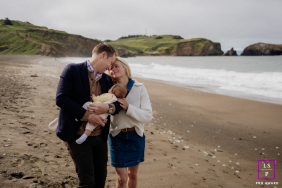In Marin, CA, mom and dad stand on the beach in gentle morning light, tenderly touching heads as they hold their newborn together, surrounded by the tranquil seaside atmosphere.