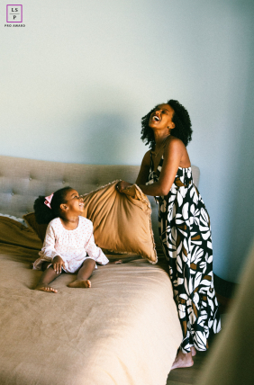 In Castro Valley, California, a young girl sits on a bed laughing joyfully while her mother stands beside her. Their shared laughter captures the warmth and happiness of childhood together.