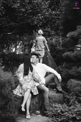 In a black and white vertical portrait at Shinn Historical Park and Arboretum, a boy stands behind his mom and dad in nature, playfully mimicking their kiss with a joyful expression.