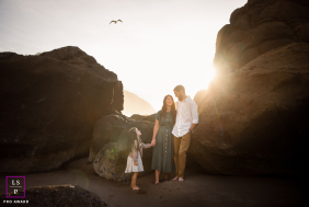 Oregon Coast Sunset: Family Smiles Between Rocky Formations as Gull Soars In Portland, Oregon, a family smiles at each other during a coastal sunset, with rocky formations behind them and a gull soaring overhead.