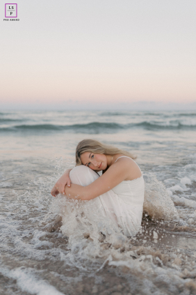 In Sheboygan, Wisconsin, a smiling teen girl sits in the water at sunset, surrounded by gentle waves and a sky filled with soft pastel colors.