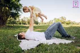 Father Lifts Daughter Joyfully Into Air In Lyon Park In Lyon, France, dad lies on his back on a blanket among grass and trees, lifting his daughter into the air with a joyful expression.