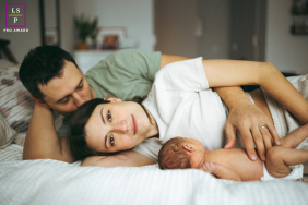 New York City Newborn Session: Cozy Family Of Three At Home In New York City, a family of three lies together on a bed at home during a newborn session, capturing the coziness of in-home life.