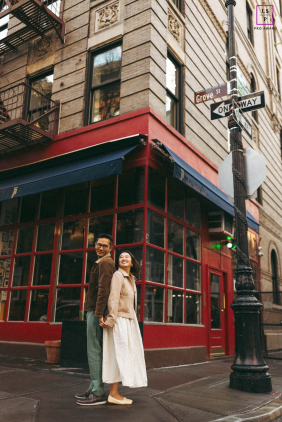 Outside Little Owl in West Village, Manhattan, a couple stands back to back, both smiling, framed by the lively city street.