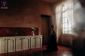 A serene woman is seen indoors, softly looking out through a large window.