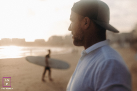 A man stands on a Vendée beach as a surfer walks past with a board in the background.