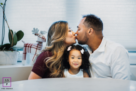 San Mateo Sun-Kissed Family: A Toddler's Table View of Parental Love A family portrait in San Mateo, California, shows a toddler sitting at a table looking at the camera as parents sit beside her, kissing over her head.