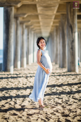 A maternity portrait in Huntington Beach, California, shows a solo woman standing below the pier between pilons in low sunlight.