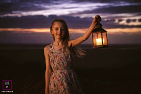 The girl holds her lantern high, casting a warm, defiant glow against the bruised purple of the Lancashire twilight. This centered composition creates a wonderful mystery, turning her into a timeless, dramatic guardian of the dunes.