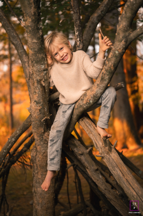 A barefoot child perches within a wooden throne, his grin capturing pure mischief. Golden light wraps the scene, turning a simple tree climb into a timeless, masterfully composed story of joy.