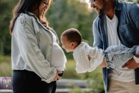 In Toronto, a father playfully lifts his daughter toward her mother’s pregnant belly as the child leans forward with excitement. The soft outdoor light and natural interaction give the photograph warmth, humor, and joyful anticipation.