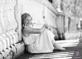 A young girl in a white dress sits barefoot beside a decorative bridge, smiling during a quiet rest in Blackpool, Lancashire.