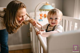 Smiling Baby Holds Hands With Mom While Standing In Connecticut Crib A smiling baby stands inside a crib while playfully holding hands with mom during a joyful family portrait in Connecticut.