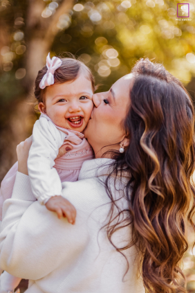 Mother Kisses Smiling Daughter During Outdoor Family Time In Curitiba Brazil A mother kisses her smiling young daughter on the cheek during a warm outdoor family lifestyle session in Curitiba, Paraná, Brazil.