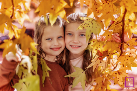 Two Sisters Smile Together Among Colorful Autumn Leaves In Curitiba Brazil Two sisters smile together while surrounded by colorful autumn leaves during a playful outdoor family session in Curitiba, Paraná, Brazil.