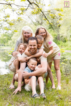 Playful Family Piles Together With Laughter Beside Pond In Overijssel A playful family piles together with laughter beside a peaceful pond during a relaxed outdoor lifestyle session in Overijssel, Netherlands.