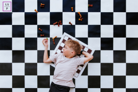 A young boy lies across a giant chessboard surrounded by scattered pieces during a playful lifestyle session in Biarritz, France.