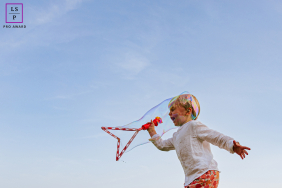 A young child runs beneath a giant floating soap bubble, surrounded by open blue sky during playful outdoor fun in Orthez, France.