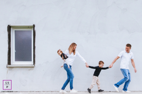 Family Walks Hand In Hand Beside White Wall In Bardos France A family walks playfully together while holding hands beside a simple white wall during a lifestyle session in Bardos, Pyrénées-Atlantiques, France.