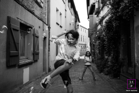 Children Chase Floating Soap Bubbles Along Cobblestone Street In Haute-Garonne Children chase and reach for floating soap bubbles while playing together along a narrow cobblestone street in Haute-Garonne, France.