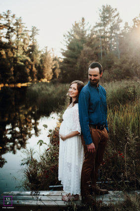 An expectant couple stands beside a quiet stream, with the mother gently cradling her pregnant belly in Braeside, Ontario, Canada.