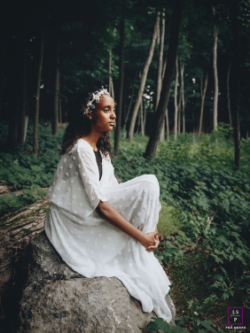 A young woman in a flowing white dress sits quietly on a fallen tree surrounded by dense forest in Braeside, Ontario, Canada.