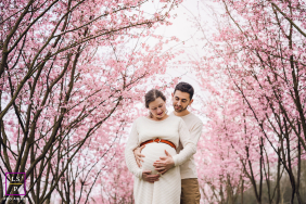 An expectant couple gently cradles her pregnant belly beneath blooming pink orchard trees during a spring maternity session in Lyon, France.
