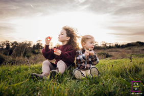 Two Children Blow Bubbles Together In Grassy Field In Lyon France Two young children sit together in a grassy field blowing bubbles as the wind sweeps through their hair in Lyon, France.