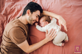 Smiling Father Gently Holds Newborn Baby Close In Lyon France A smiling father lies beside his newborn baby, gently holding them close during a lifestyle family session in Lyon, France.