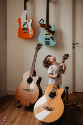 A young boy stands among towering acoustic and electric guitars, looking upward at the instruments surrounding him at home in Bordeaux, France.