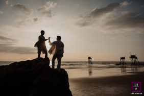 A couple playfully connects in silhouette atop seaside rocks as the sun sets over the beach and ocean in Pornic, France.