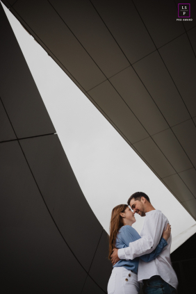 A couple embraces beneath dramatic geometric architecture, sharing a quiet affectionate portrait framed by sweeping lines in Angers, France.