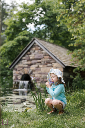 A young girl gently smells a flower beside a peaceful garden pond and small stone cottage in New Hope, Pennsylvania.