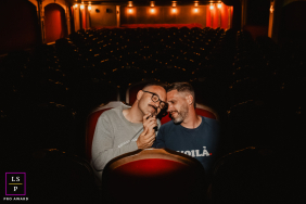 A couple cuddles closely in theater seats, smiling together amid the warm glow and empty rows of a theater in Nantes, France.