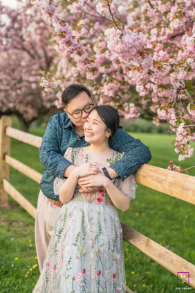A couple shares a joyful embrace beneath blooming pink trees, smiling together during a springtime session in Flemington, New Jersey.