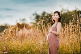 An expectant mother gently cradles her pregnant belly while smiling among tall grasses in warm sunset light in Far Hills, New Jersey.