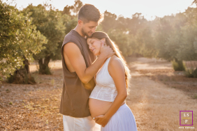 An expectant couple embraces tenderly in warm sunlight, gently cradling her pregnant belly among the olive groves of Jaén, Spain.